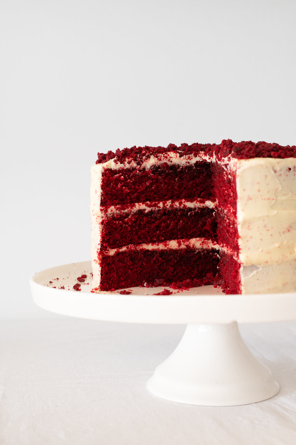 A close up of a Red Velvet Cake covered in cream cheese icing with red crumb garnish on top, showing three layers