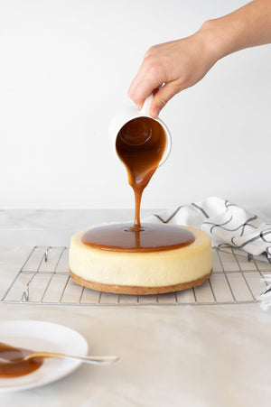 Caramel glaze being poured over a cheesecake on a cooling rack
