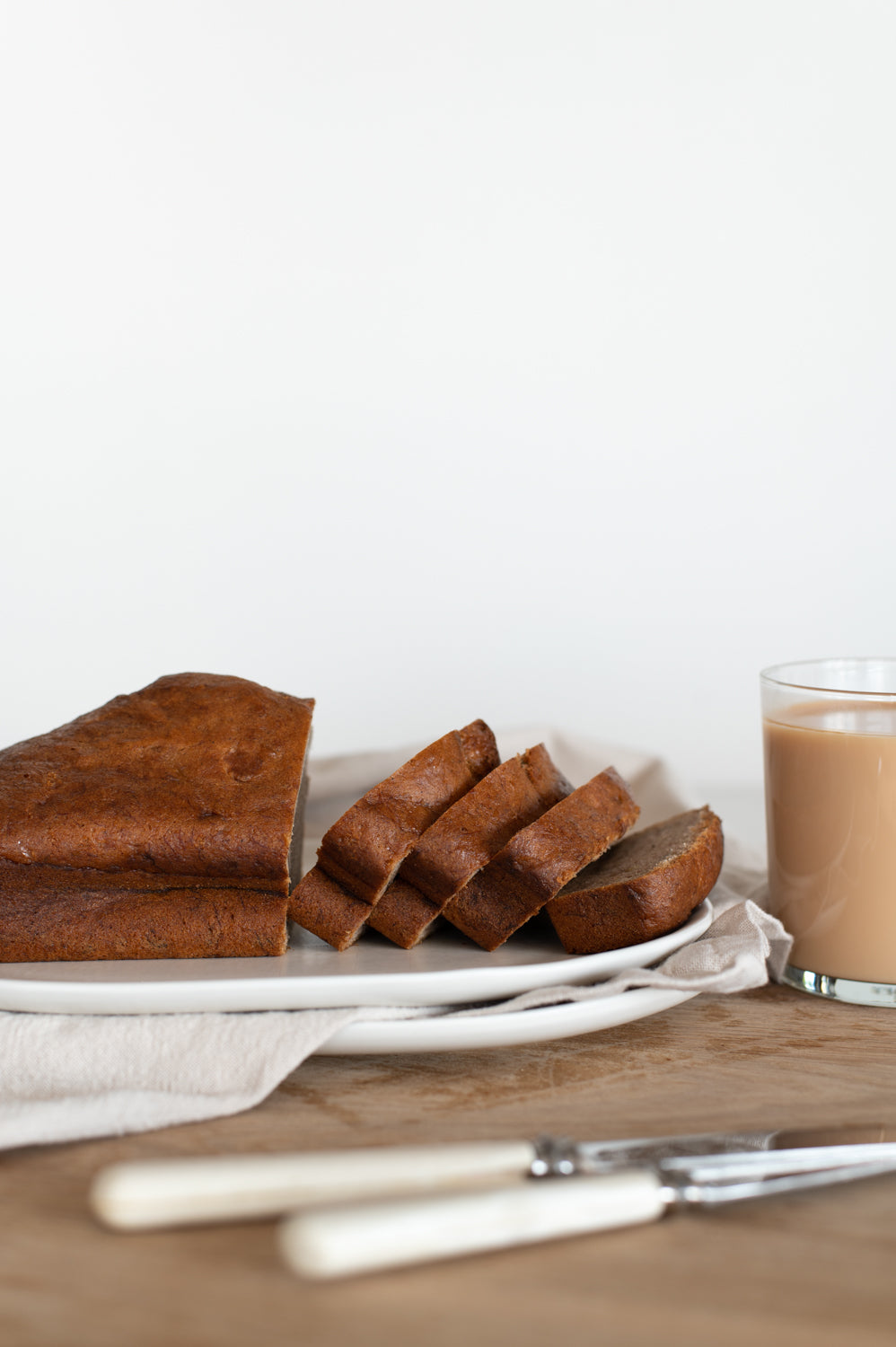 A loaf of sliced vegan banana bread on a wooden board with a glass of tea